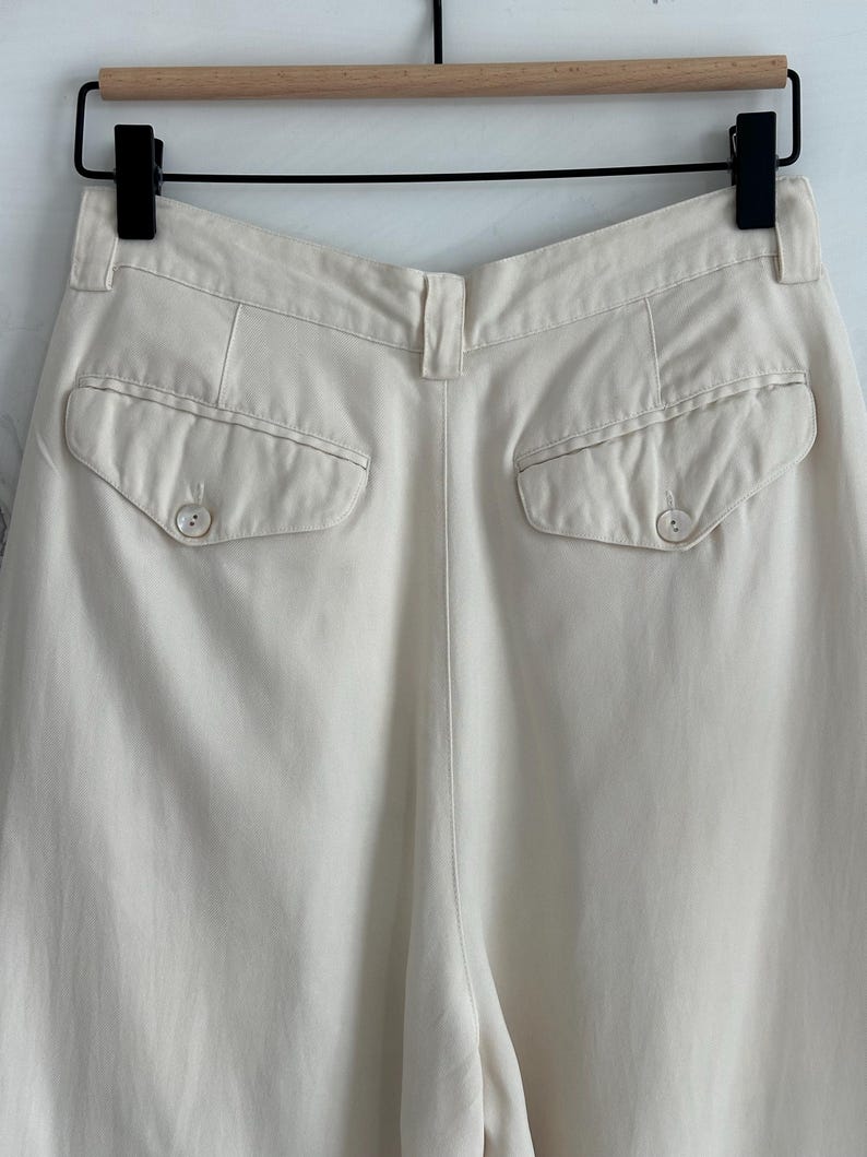 Puede incluir: Pantalones color crema con dos bolsillos traseros abotonados. Los pantalones est&aacute;n colgados de una percha negra con una barra de madera. La tela parece ser un material ligero, adecuado para climas c&aacute;lidos. Los pantalones tienen cintura alta y corte holgado.