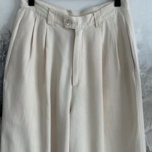 Puede incluir: Pantalones plisados ​​de color crema con cierre de bot&oacute;n y pernera recta. Los pantalones cuelgan de una percha negra sobre un fondo neutro. La tela parece ser un material ligero.