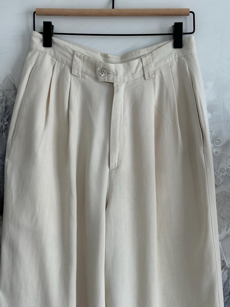 Puede incluir: Pantalones plisados ​​de color crema con cierre de bot&oacute;n y pernera recta. Los pantalones cuelgan de una percha negra sobre un fondo neutro. La tela parece ser un material ligero.