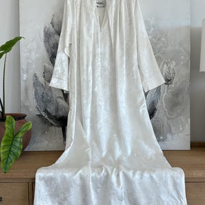 Pode incluir: Um roupão branco de manga comprida com um padrão floral jacquard tonal subtil em todo o tecido. A peça tem uma gola alta, um fecho frontal com botões e uma etiqueta na linha do pescoço com a inscrição ARIEL. A bainha vai até ao chão.