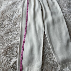 Puede incluir: Pantalones color crema de pernera ancha con la parte delantera plisada. Una cinta m&eacute;trica rosa muestra la longitud de los pantalones. Los pantalones miden aproximadamente 114 cm de largo. La tela tiene un brillo sutil. Los pantalones est&aacute;n sobre una superficie blanca y esponjosa.