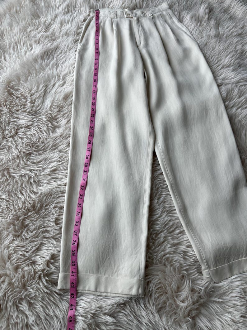 Puede incluir: Pantalones color crema de pernera ancha con la parte delantera plisada. Una cinta m&eacute;trica rosa muestra la longitud de los pantalones. Los pantalones miden aproximadamente 114 cm de largo. La tela tiene un brillo sutil. Los pantalones est&aacute;n sobre una superficie blanca y esponjosa.