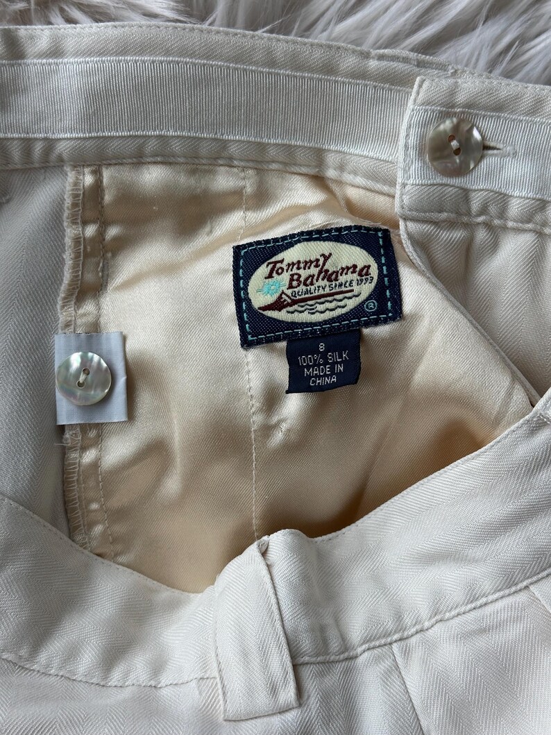 Puede incluir: Primer plano de un pantal&oacute;n de seda color crema con cierre de bot&oacute;n. La etiqueta Tommy Bahama es visible, junto con una etiqueta de talla 8. El pantal&oacute;n est&aacute; hecho de 100% seda.