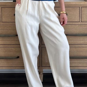 Puede incluir: Pantalones color crema de pernera ancha con cintura alta y pliegues. Los pantalones se combinan con una blusa azul marino. El conjunto se completa con una pulsera dorada y zapatos color crema de punta.