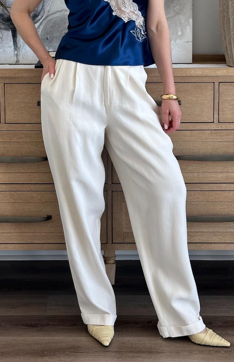 Puede incluir: Pantalones color crema de pernera ancha con cintura alta y pliegues. Los pantalones se combinan con una blusa azul marino. El conjunto se completa con una pulsera dorada y zapatos color crema de punta.