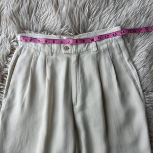 Puede incluir: Pantalones plisados color crema con cierre de bot&oacute;n y cinta m&eacute;trica rosa en la cintura. La cinta muestra medidas en pulgadas. Los pantalones son de tela lisa y est&aacute;n colocados sobre una superficie suave y clara.