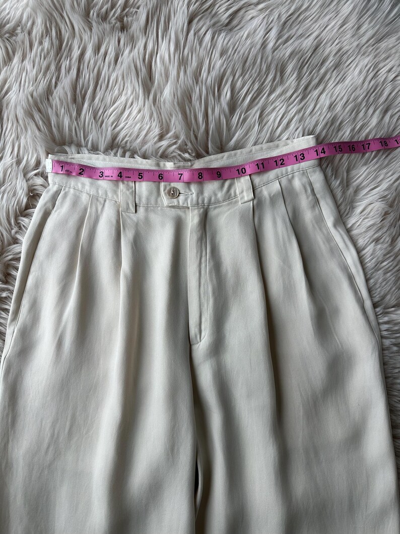 Puede incluir: Pantalones plisados color crema con cierre de bot&oacute;n y cinta m&eacute;trica rosa en la cintura. La cinta muestra medidas en pulgadas. Los pantalones son de tela lisa y est&aacute;n colocados sobre una superficie suave y clara.