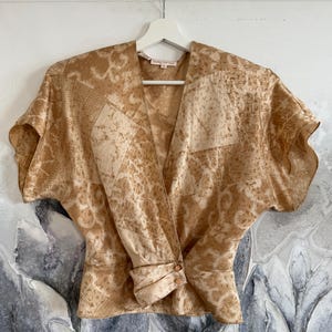Può includere: Una blusa a portafoglio a maniche corte color marrone chiaro e beige con un motivo stile patchwork caratterizzato da varie texture astratte. Il capo presenta uno scollo a V, maniche ampie e un dettaglio arricciato in vita con due bottoni.