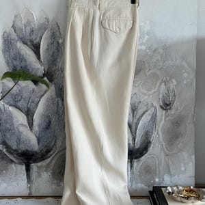 Puede incluir: Pantalones color crema de cintura alta con un bolsillo trasero abotonado y pliegues. Los pantalones cuelgan de una percha negra sobre un fondo de pintura floral. La tela parece ser ligera.