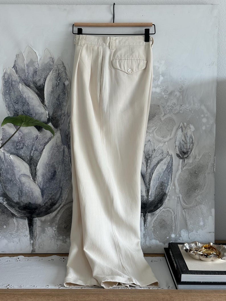 Puede incluir: Pantalones color crema de cintura alta con un bolsillo trasero abotonado y pliegues. Los pantalones cuelgan de una percha negra sobre un fondo de pintura floral. La tela parece ser ligera.