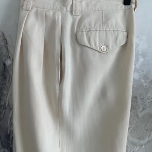 Puede incluir: Pantalones color crema con la parte delantera plisada y un bolsillo trasero con cierre de bot&oacute;n. Los pantalones est&aacute;n colgados de una percha de madera. La tela parece ser un material ligero.