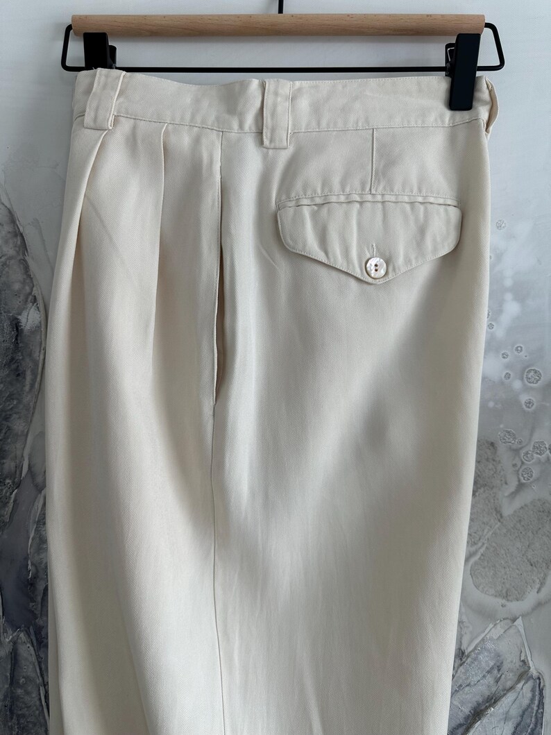 Puede incluir: Pantalones color crema con la parte delantera plisada y un bolsillo trasero con cierre de bot&oacute;n. Los pantalones est&aacute;n colgados de una percha de madera. La tela parece ser un material ligero.