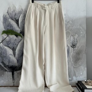 Puede incluir: Pantalones color crema de pernera ancha colgados de una percha de madera. Los pantalones tienen cierre de bot&oacute;n y pliegues en la cintura. La tela parece ser un material ligero. Los pantalones se muestran contra una pintura floral.