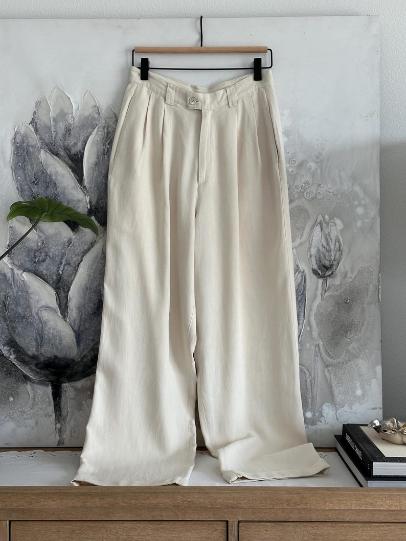 Puede incluir: Pantalones color crema de pernera ancha colgados de una percha de madera. Los pantalones tienen cierre de bot&oacute;n y pliegues en la cintura. La tela parece ser un material ligero. Los pantalones se muestran contra una pintura floral.