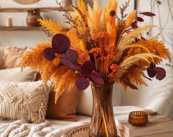 Peut inclure: Un arrangement décoratif d'herbes de la pampa séchées, de plumes orange et de feuilles d'eucalyptus bordeaux foncé dans un vase en verre ambré cannelé. Le bouquet présente des textures variées et des tons automnaux chauds.