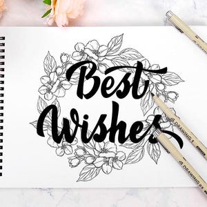 May include: A spiral-bound sketchbook with the words "Best Wishes" in elegant script, surrounded by a floral wreath. Several beige drawing pens are scattered around the sketchbook, with peach-colored flowers in the background.