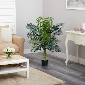 May include: A lush, artificial palm tree with vibrant green fronds and a slender, light brown trunk. The plant is potted in a dark green container and stands on a gray wood-look floor. The scene includes a beige sofa, a white coffee table, and a decorative side table.