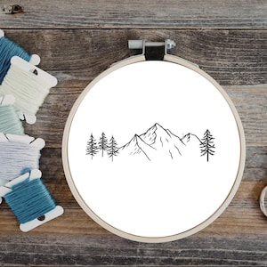 Può includere: Un telaio da ricamo con un design minimalista di montagne e alberi in filo nero. Varie bobine di filo da ricamo colorato e un paio di forbici vintage sono disposte su una superficie di legno, suggerendo un progetto artigianale.