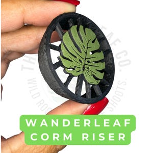May include: A circular black plant support structure featuring a textured outer rim and internal spokes radiating from a central olive green monstera leaf design. The text WANDERLEAF CORM RISER is printed in white on a green rectangular banner.
