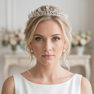 Könnte beinhalten: Ein silberfarbenes Diadem mit einer symmetrischen Anordnung aus klaren, facettierten Kristallen in Blatt- und Tropfenform. Das Metallband ist detailliert mit kleinen, funkelnden Steinen verziert, was ein filigranes, erhöhtes Kronendesign ergibt.