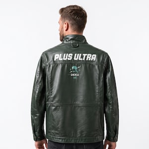 以下が含まれることがあります： スタンドカラーと長袖のダークグリーンのレザージャケット。背面には「PLUS ULTRA」の白い文字がプリントされており、その下にはデクのシルエットのグラフィックと「DEKU」、括弧で囲まれた「A」の文字がデザインされています。