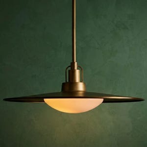 May include: A pendant light fixture featuring a wide, flat, circular metal shade with a brushed brass finish. A central, semi-spherical frosted glass diffuser is positioned beneath the shade, emitting a soft, warm glow from the light source.