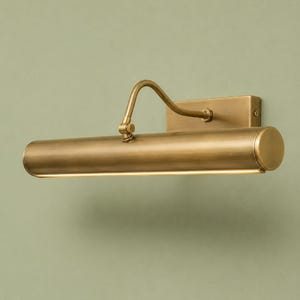 May include: A wall-mounted picture light with a brushed brass finish. The fixture features a rectangular backplate, a curved adjustable arm, and a long cylindrical lamp head with a thin horizontal light opening along the bottom edge.