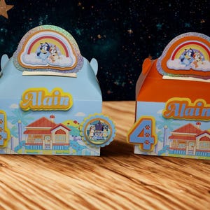 May include: Two colorful party favor boxes, one blue and one orange, with a cartoon theme. Each box features the name "Alain", the number "4", and a cartoon house illustration. The boxes are decorated with a rainbow and character images.