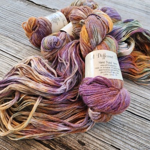 May include: A Different Twist hand-dyed yarn in a mix of purple, orange, and brown shades. The yarn is wound into a ball and is ready to be used for knitting or crocheting.