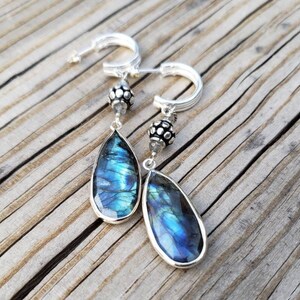 May include: A pair of silver hoop earrings with teardrop-shaped labradorite stones. The stones have a blue and green iridescent sheen.