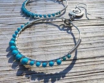 May include: A pair of silver hoop earrings with turquoise blue beads. The earrings have a spiral design at the top and are set against a weathered wooden surface. The beads are evenly spaced along the bottom half of the hoops.