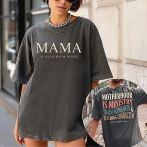 May include: Dark gray t-shirt with the word "MAMA" above "IS KINGDOM WORK" in a serif font. The back of the shirt has the text "MOTHERHOOD IS MINISTRY TRAINING HEARTS & BUILDING CHARACTER" and "PROVERBS 22:6".