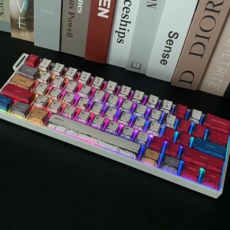 May include: A white mechanical keyboard with a variety of colourful keycaps. The keys feature a cartoon design and are in shades of red, blue, yellow, and white. The keyboard has a white frame and sits on a black surface.