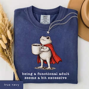 Könnte beinhalten: Marineblaues T-Shirt mit einem Cartoon-Frosch in einem roten Umhang, der eine Kaffeetasse hält. Der Text auf dem Shirt lautet "being a functional adult seems a bit excessive". Die Marke des Shirts ist Comfort Colors.
