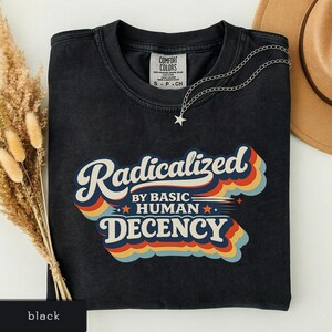 May include: Black t-shirt with the text "Radicalized by Basic Human Decency" in a retro font. The design features a layered, colorful design with blue, orange, and yellow accents. A silver necklace with a star pendant is draped on the shirt.