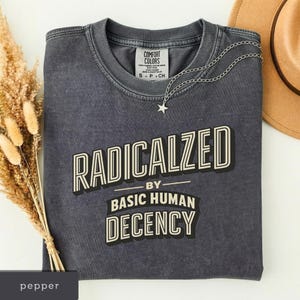 Può includere: Una maglietta grigio scuro con la scritta "RADICALIZED BY BASIC HUMAN DECENCY" in grassetto e contornata. Una collana d'argento con un ciondolo a stella è appoggiata sulla maglietta piegata, con un'etichetta visibile.