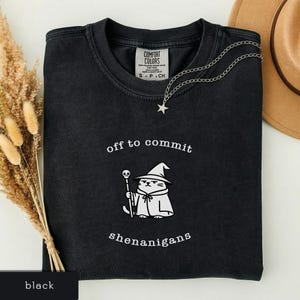 Puede incluir: Camiseta negra con la frase "off to commit shenanigans" y un gato de dibujos animados disfrazado de mago. El gato lleva un sombrero de mago y sostiene un bastón. Un collar plateado con un colgante de estrella está sobre la camiseta.