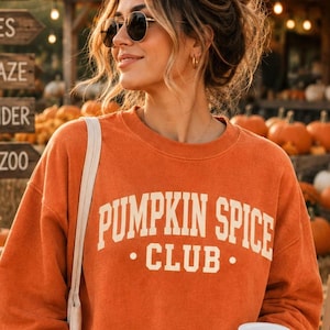 May include: An orange crewneck sweatshirt featuring the text PUMPKIN SPICE CLUB in cream-colored block lettering across the chest. The long-sleeved garment is worn with a white tote bag and a white paper coffee cup with a lid.