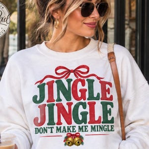 May include: A cream-colored long-sleeve sweatshirt featuring a red and green graphic print that reads JINGLE JINGLE DON'T MAKE ME MINGLE with a red bow at the top and two gold jingle bells at the bottom, paired with blue denim jeans.