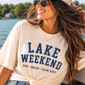 K&ouml;nnte beinhalten: Ein cremefarbenes Kurzarm-T-Shirt mit dunkelblauem Text, der LAKE WEEKEND SUN WATER SLOW DAYS lautet. Das Shirt wird mit einer hellblauen Jeansshorts, einer marineblau-beigen Trucker-Cap und einer goldfarbenen Halskette mit Anh&auml;nger kombiniert.
