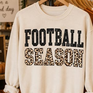 May include: A cream-colored crewneck sweatshirt featuring the text FOOTBALL SEASON printed on the front. The word FOOTBALL is in black, and the word SEASON is in a brown and black leopard print pattern. The sweatshirt has long sleeves and a ribbed collar.