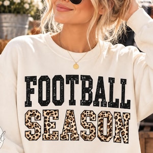 May include: A cream-colored crewneck sweatshirt featuring the text FOOTBALL SEASON printed on the front. The word FOOTBALL is in a distressed black font, while the word SEASON is in a black-outlined font filled with a brown leopard print pattern.