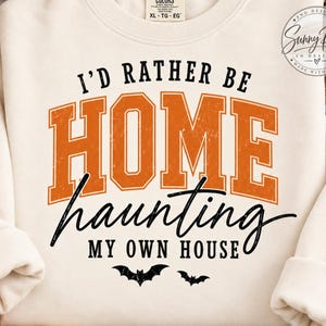 Puede incluir: Una sudadera de cuello redondo color crema con el texto I'D RATHER BE HOME haunting MY OWN HOUSE en letras naranjas y negras. El diseño incluye dos pequeñas siluetas de murciélagos negros en la parte inferior y una etiqueta Comfort Colors.