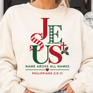 May include: A cream-colored crewneck sweatshirt featuring a graphic print that spells JESUS in red and green patterns. Below the text, it reads NAME ABOVE ALL NAMES, a small red heart, and the scripture reference PHILIPPIANS 2:9-11 in red text.