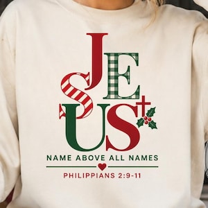 May include: A cream-colored crewneck sweatshirt featuring a graphic print that reads JESUS in red and green, with the text NAME ABOVE ALL NAMES and PHILIPPIANS 2:9-11 below. The design includes a small cross, holly leaves, and a red heart.