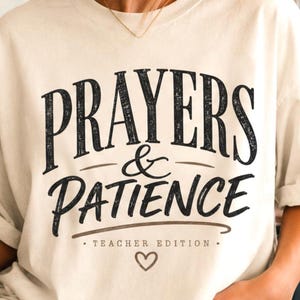 May include: A cream-colored short-sleeve t-shirt featuring black text that reads PRAYERS & PATIENCE TEACHER EDITION with a small heart symbol below the text. The shirt has a crew neckline and a relaxed fit, worn with light blue denim jeans.