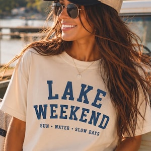 K&ouml;nnte beinhalten: Ein cremefarbenes Kurzarm-T-Shirt mit dem dunkelblauen Schriftzug LAKE WEEKEND in fetter Schrift und dem kleineren Text SUN WATER SLOW DAYS darunter. Das T-Shirt hat einen Rundhalsausschnitt und eine lockere Passform aus baumwollartigem Stoff.