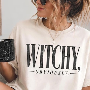 Może przedstawiać: Kremowy t-shirt z krótkim rękawem z czarnym napisem "WITCHY, OBVIOUSLY." nadrukowanym z przodu. Koszulka ma okrągły dekolt i luźny krój, a grafika jest wyśrodkowana i wykonana pogrubioną czcionką szeryfową o lekko postarzanej teksturze.