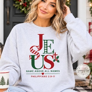 May include: A light gray crewneck sweatshirt featuring the word JESUS in red and green, with a plaid pattern on the letter E. Below the text is the phrase NAME ABOVE ALL NAMES, a small red heart, and the scripture reference PHILIPPIANS 2:9-11.
