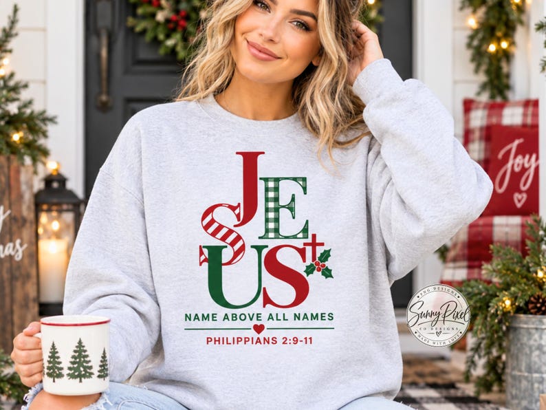 May include: A light gray crewneck sweatshirt featuring the word JESUS in red and green, with a plaid pattern on the letter E. Below the text is the phrase NAME ABOVE ALL NAMES, a small red heart, and the scripture reference PHILIPPIANS 2:9-11.